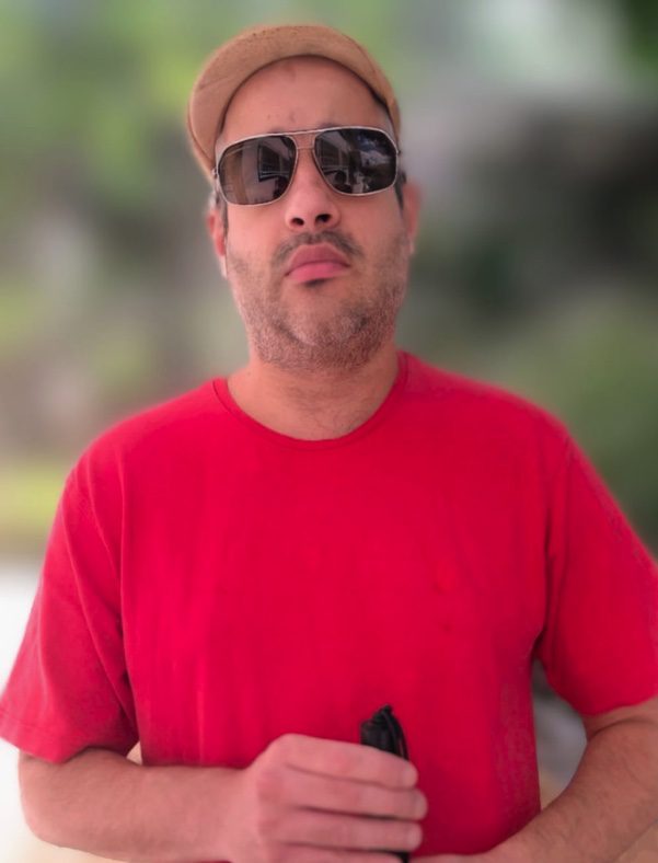 Na imagem temos Manoel Negraes sorrindo, ele está usando camiseta vermelha, seus cabelos estão levemente bagunçados, ao fundo uma sala com quadros diversos nas paredes. 